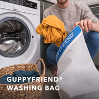 Person using a Guppyfriend washing bag with a washing machine and laundry basket in the background.