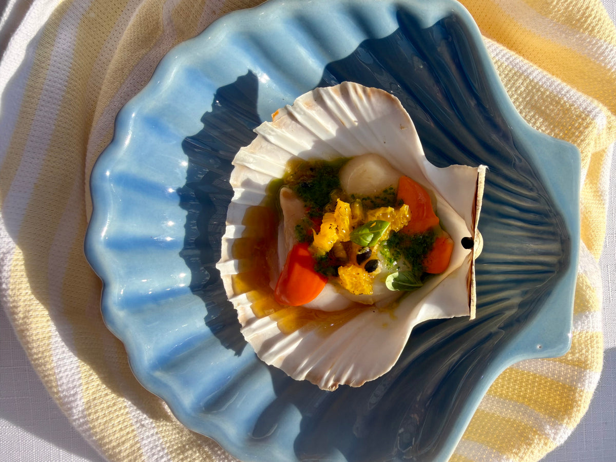 A scallop shell filled with cooked seafood and vegetables, presented on a blue scallop-shaped plate over a yellow-and-white striped cloth.